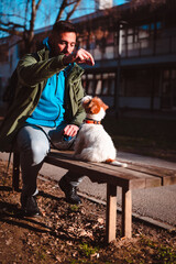 Young man training his Jack Russell terrier puppy outdoor in park. Cute little doggie listening to commands from its owner and waiting for treats.