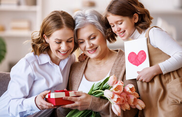 Happy International Women's Day.Smiling  daughter and granddaughter giving flowers  and gift to grandmother   celebrate spring holiday Mother's Day at home - 490338935