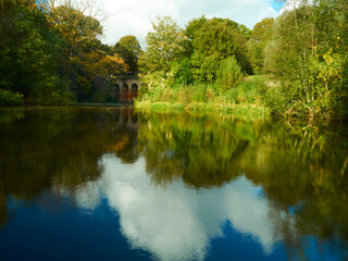 Fototapeta na wymiar Rich, saturated, sunlit view of Hampstead Viaduct with the structure, trees and deep blue sky reflected in still lake water.