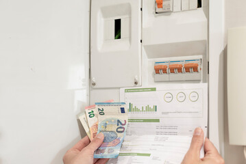 a hand holding the electricity bill and euro banknotes in front of power control switch