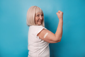 Vaccinated 50s mature older senior woman getting a vaccine protection the coronavirus. Happy female showing arm with bandage after receiving vaccination over blue background. 2024 - obrazy, fototapety, plakaty