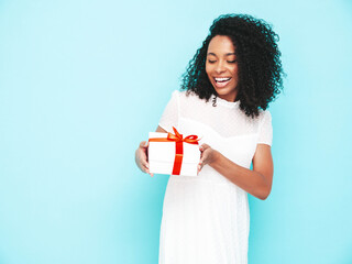 Beautiful black woman with afro curls hairstyle. Smiling model dressed in white summer dress. Sexy carefree female posing near blue wall in studio. Tanned and cheerful. Holding gift box. Isolated