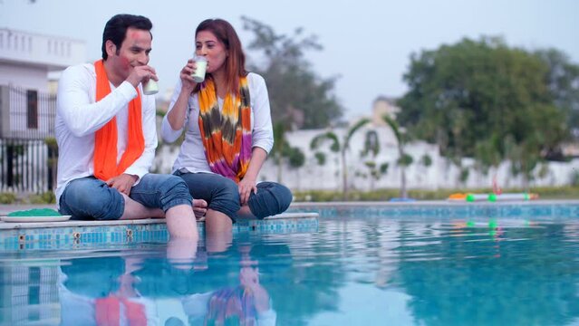 A young Indian couple smeared with Holi colors on their faces drinking Thandie together - happy faces togetherness and bonding cheerful. Office colleagues/friends wearing white Kurta and a Dupo