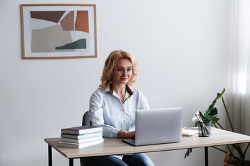 Portrait of adult blonde woman wearing blue blouse and glasses sitting at her workplace. Smart looking businesswoman working on a laptop in the office. Background, copy space, close up.