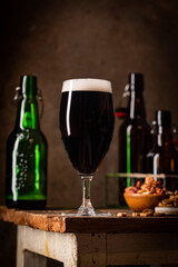Glass of dark beer with foam head on dark wooden background with empty bottles and beer snacks