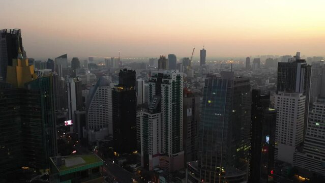 Drone descending above skyline and skyscrapers of Sukhumvit district during colorful warm sunrise in Bangkok, Thailand.