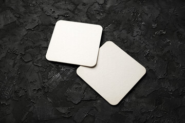 Two blank square beer coasters on black stone background.