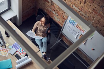 High angle view of female interior designer looking aside, drinking coffee or tea, holding a blueprint while working on new project, sitting at workplace