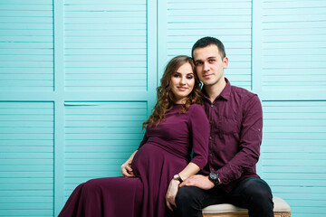 A pregnant brunette and a happy husband are sitting on a sofa looking into the distance, happy motherhood. Turquoise wall and gray soft armchairs.