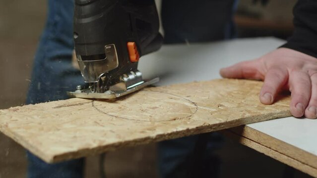 Artisan holds an osb board i craft room and cuts with jig saw in circle, front view. Craftsman works with osp saw it with fretsaw