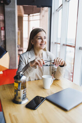 A young woman manager in a light jacket and glasses, a girl with a smile on her face, sits at a wooden table with a laptop and a phone. Remote work