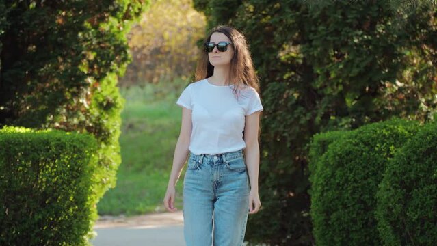 A beautiful young brunette woman in sunglasses is walking through a summer park among green trees.