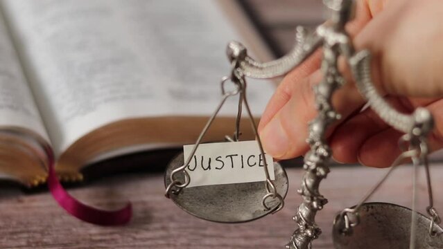 Hand holding a handwritten text "justice" and leaving it on an old-fashioned silver weighing balance scale with open Holy Bible Book in the background. A closeup. The concept of Law and righteousness.