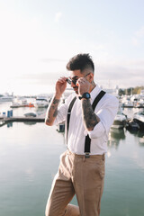 A handsome stylish brutal man a multiracial American Spaniard with a fashionable beard and sunglasses in a wedding suit against the background of the sea and the port with yachts and boats outdoors 