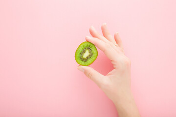 Young woman fingers holding half of fresh green kiwi on light pink table background. Pastel color. Closeup. Top down view.