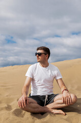 Fototapeta na wymiar Man in sunglasses sitting in lotus pose in desert on sand