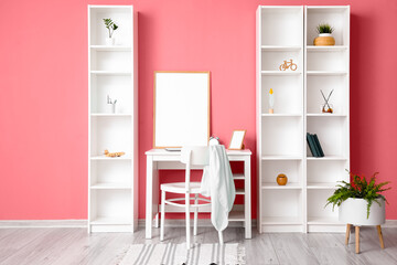 Fototapeta na wymiar Modern workplace with blank frame and shelving units near pink wall