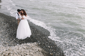 Fototapeta na wymiar The bride and groom in wedding dresses hug and walk together along the sea in nature on an outdoor trip. Husband and wife in love spend their honeymoon. Selective focus