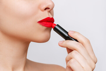 Cropped shot of a young beautiful caucasian woman applying a bright red color lipstick on her lips isolated on a gray background. Close-up of using lipstick, make up