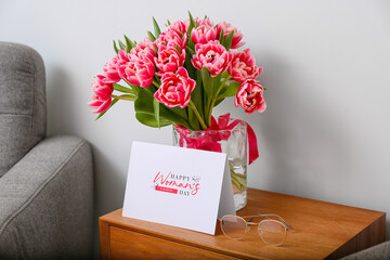 Tulips, eyeglasses and greeting card with text HAPPY WOMAN'S DAY on table near light wall