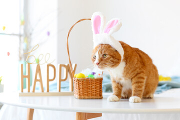 Cute cat with bunny ears, basket and Easter eggs on table in bedroom