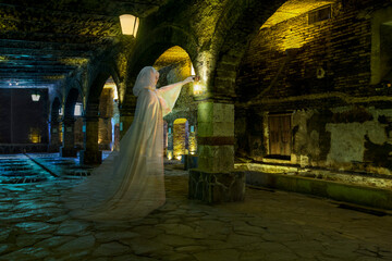 Female ghost with lantern, dressed all in white standing in a cavernous stone basement 