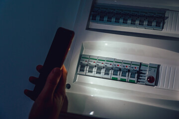 Energy crisis. Hand in complete darkness holding a phone to investigate a home fuse box during a...