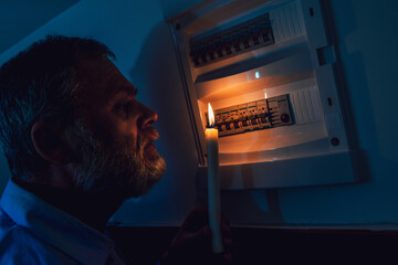 Energy crisis. Man in complete darkness holding a candle to investigate a home fuse box during a...