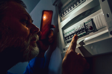 Energy crisis. Man in complete darkness holding a phone to investigate a home fuse box during a...