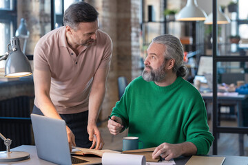 Mature bearded businessman sitting at his workplace with laptop and talking to his colleague, they planning work in team