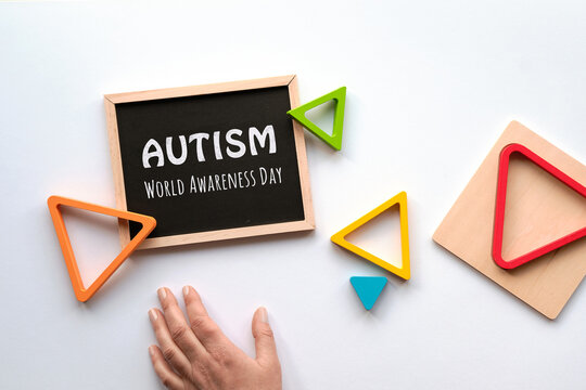 Text Autism awareness month on blackboard. Nested wood triangles in rainbow colors, human hand. Geometric education stacking puzzle. Montessori sensory toy. Flat lay on white paper background.