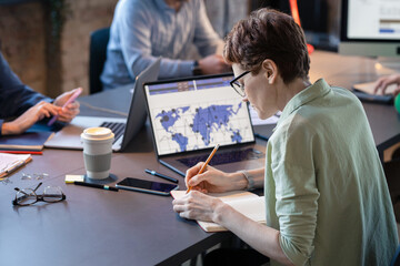 Rear view of businesswoman in eyeglasses sitting at the table with her colleagues and making notes in notebook during meeting at office