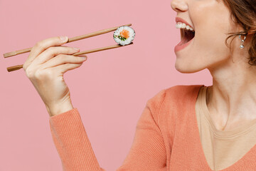 Side view close up cropped young fun woman 20s in casual clothes hold in hand eat biting with chopsticks makizushi sushi philadelphia roll japanese food isolated on plain pastel pink color background