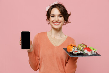 Young fun woman in casual clothes hold makizushi sushi roll served on black plate traditional japanese food use mobile cell phone blank screen workspace area isolated on plain pastel pink background