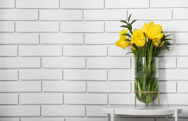 Vase with yellow tulips on table near white brick wall