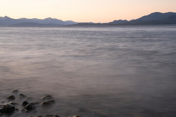 Long exposure shot of Nahuel Huapi lake at sunset. Beautiful blurred water effect, the rocky shore, mountains in the horizon and dusk colors.