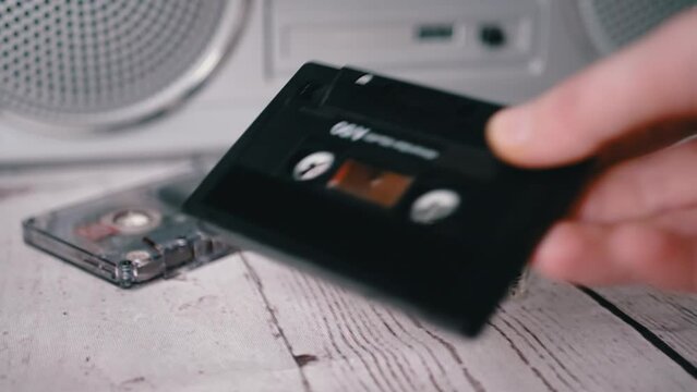 Female Hand Picks Up One Old Music Audio Cassette from the 90s from the Table. A pile of vintage magnetic tape cassettes lie on a table against the background of a stereo tape recorder. Nostalgia.