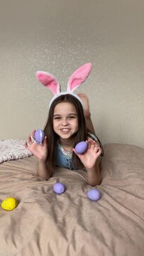 a little girl with dark hair in hare ears lies on the bed and holds purple easter eggs in her hands, happy easter.