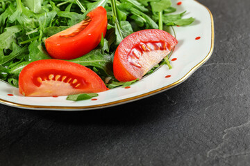 Plate with green leaves salad - arugula rocket - with fresh red tomatos on black stone desk, closeup detail