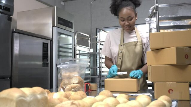 A happy African female baker in apron and gloves standing at a bakery kitchen, pasting shipping labels on parcel boxes of breads that ordered online from customers and prepared for delivery by post.