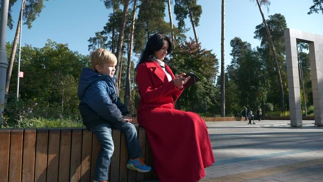 Mother and Son Watch Media on Phone. Woman Surfing Browsing Internet Using Smartphone. Mom with Kid rest in City Park. Autumn Day. 2x Slow motion 60fps 4K