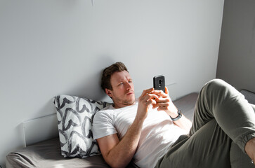 Caucasian attractive young man in white t-shirt woke up chatting, surfing, working on pc and making phone call on bed in bedroom interior. Modern device for remote communication