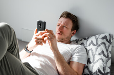 Caucasian attractive young man in white t-shirt woke up chatting, surfing, working on pc and making phone call on bed in bedroom interior. Modern device for remote communication