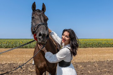 Cheerful brunette with red lipstick standing in agriculture field with brown horse. Sunny day, clear sky and sunflower field in background.