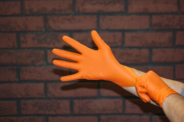 The hands of a man wearing rubber gloves are insulation against a brick wall. Close-up of orange latex protective gloves against a brick wall.