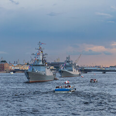 small boats with tourists inspect warships on the Neva in the city center against the blue evening sky