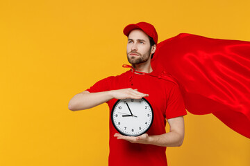 Professional minded delivery guy employee man 20s in red cap T-shirt uniform red cloak workwear work as dealer courier hold clock isolated on plain yellow background studio portrait. Service concept. - obrazy, fototapety, plakaty