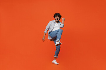 Fototapeta na wymiar Full size body length jubilant exultant young bearded Indian man 20s years old wears blue shirt do winner gesture celebrate clenching fists say yes isolated on plain orange background studio portrait.