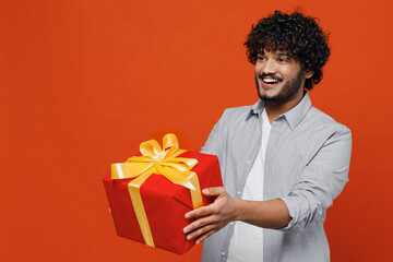 Fascinating cheerful overjoyed fun charming young bearded Indian man 20s years old wears blue shirt hold give red present box with gift ribbon bow isolated on plain orange background studio portrait. - obrazy, fototapety, plakaty