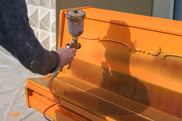 A Man's hand holding a paint spray gun and coloring a piano in an orange color